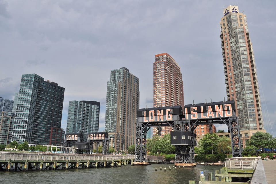 The two historic gantries at Gantry Plaza State Park in Long Island City, each carrying the orange LONG ISLAND lettering, with the East River and the LIC towers behind.
